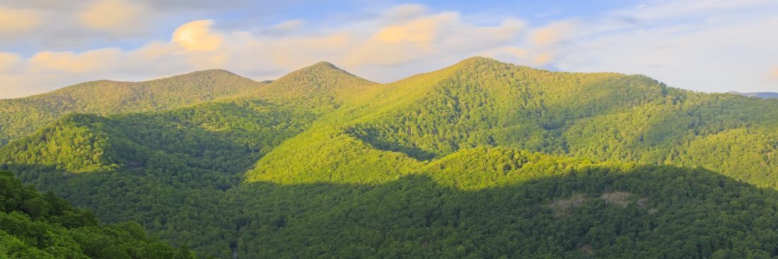 Warm evening light sweeping across overlapping green Blue Ridge summits below pastel clouds