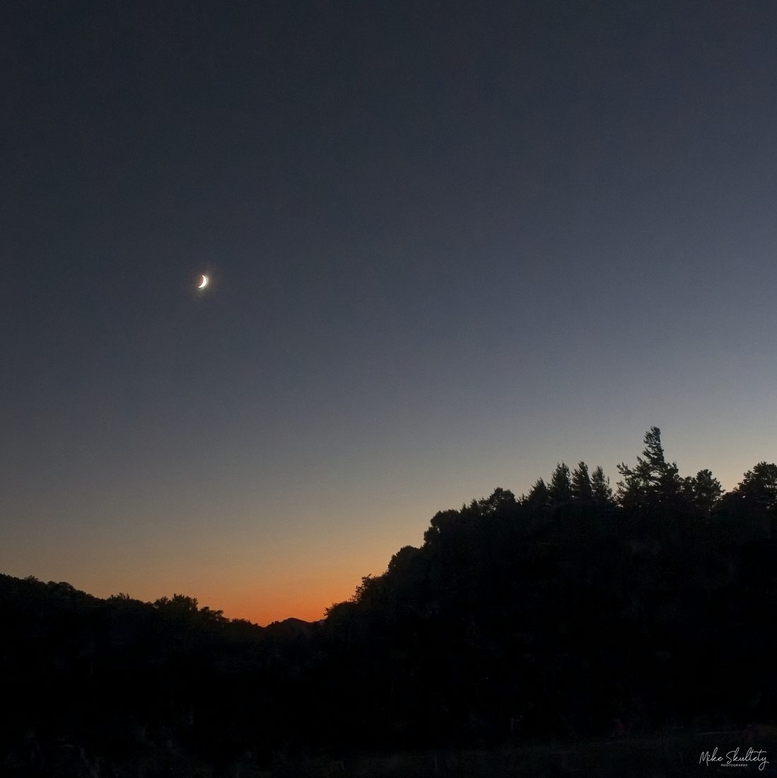 Crescent moon rising above silhouetted ridge at the blue hour